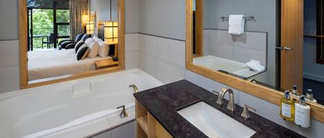Une salle de bain moderne dotée d’un grand miroir, d’une baignoire, d’un lavabo et offrant une vue sur une chambre à coucher avec un lit et un balcon.