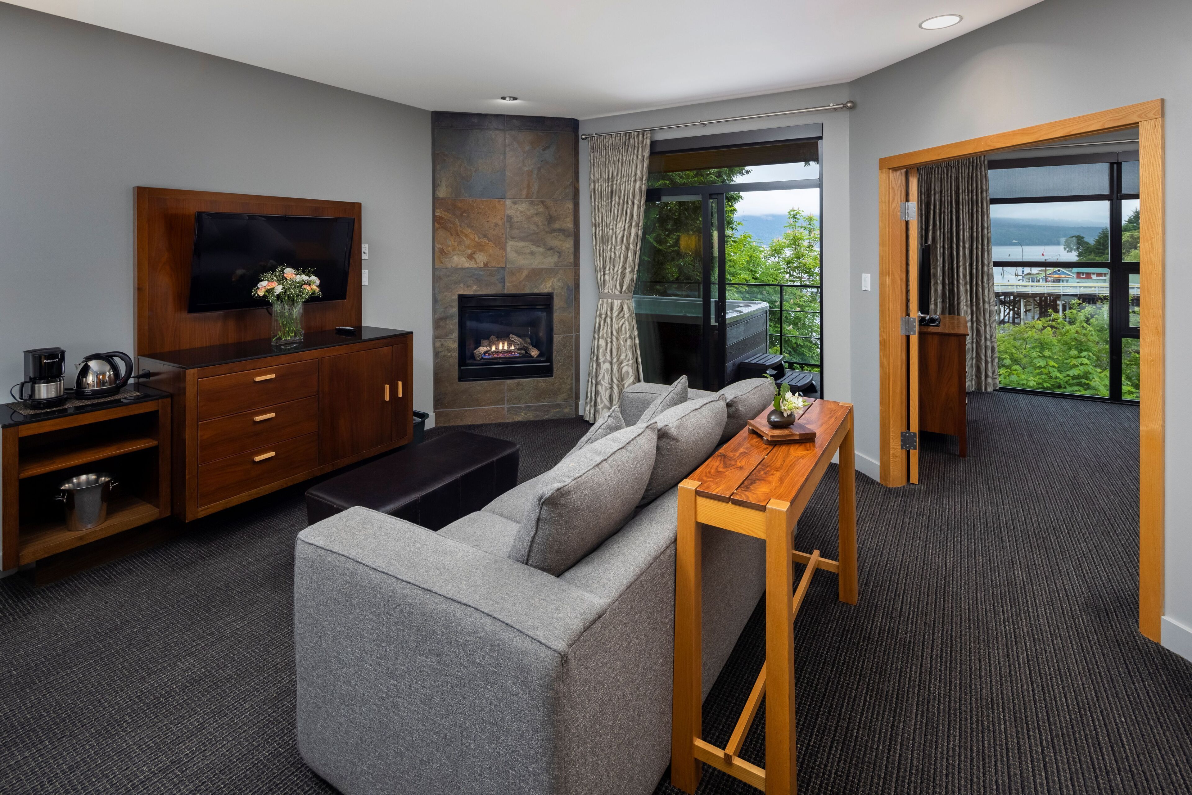 A modern living room with a grey sofa, a flat-screen TV, a fireplace, and a balcony with a view of the sea.