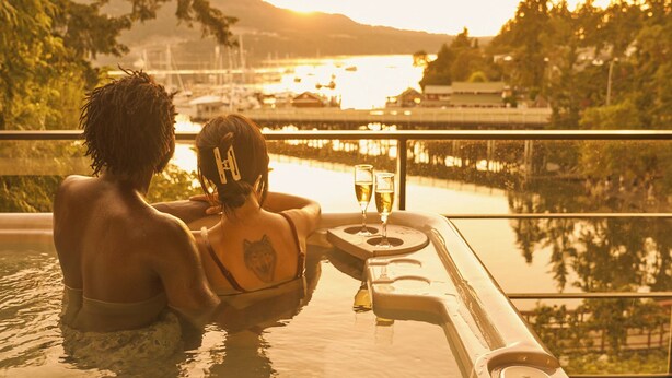 Twee personen ontspannen in een jacuzzi met uitzicht op een jachthaven en bergen.