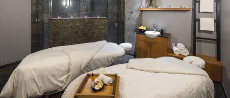 Ein moderner Spa-Raum mit zwei Massageliegen, einem großen Spiegel, einem Waschbecken und einer Holzeinrichtung.