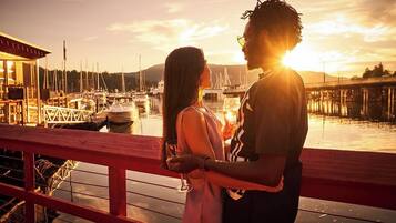 Um casal em um deck de madeira com vista para uma marina, com barcos e uma ponte ao fundo.