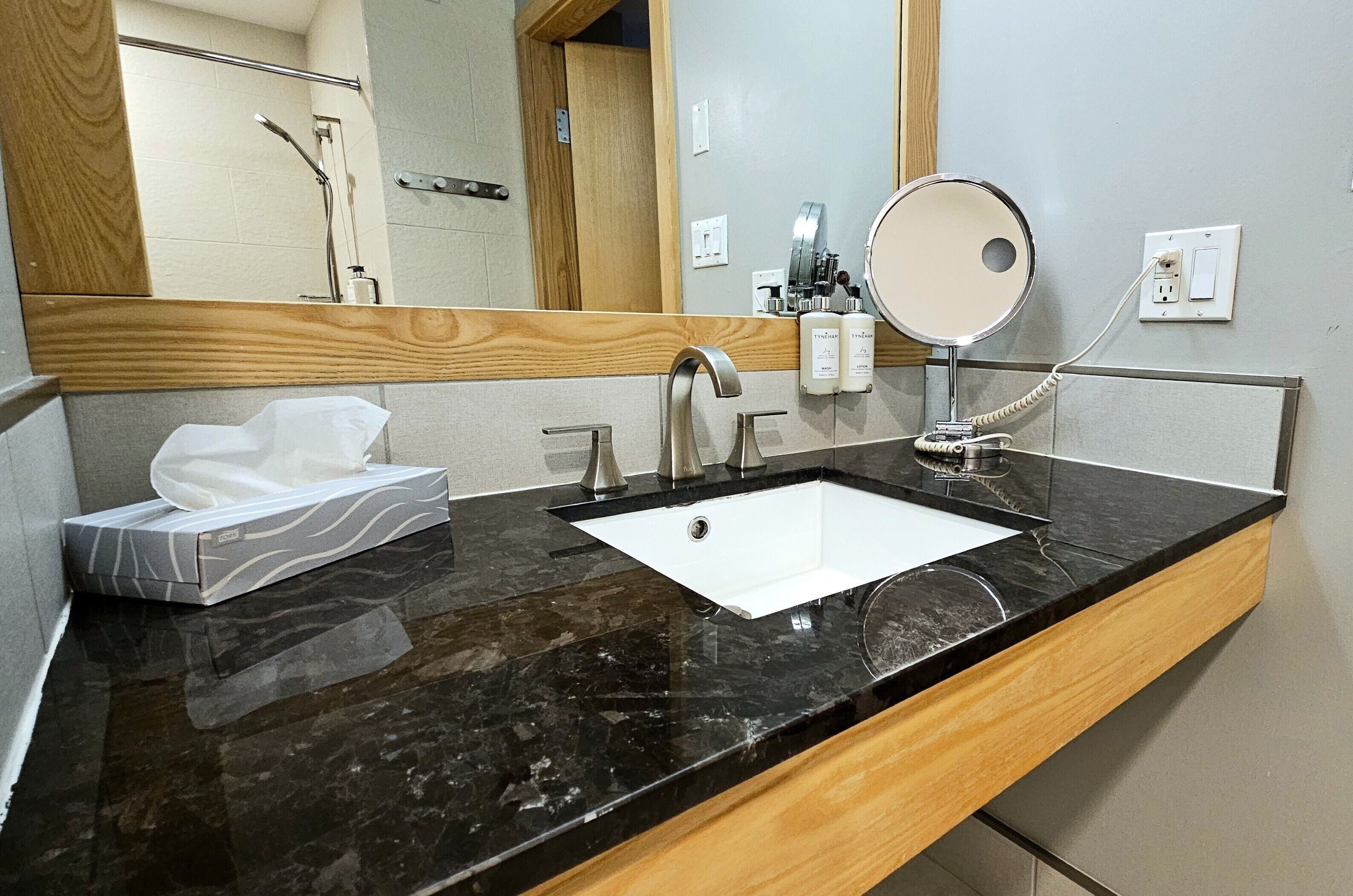A bathroom with a black marble sink, a wooden vanity, a mirror, and a shower area visible in the background.