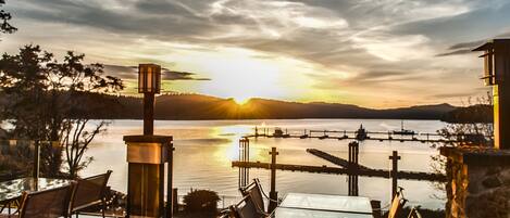 A sunset view from an outdoor dining area with a glass table and chairs, overlooking a calm lake and a distant bridge.