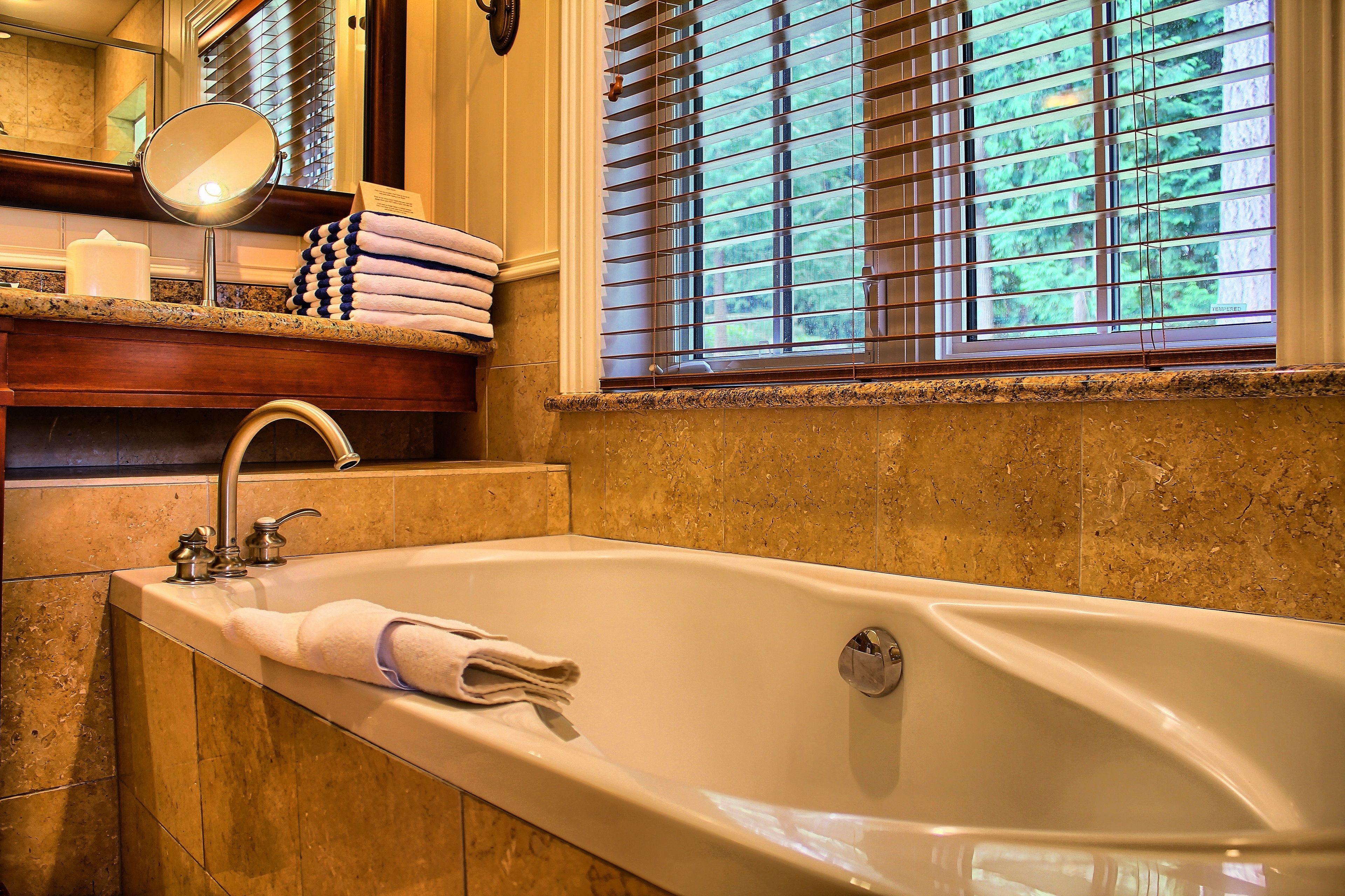A bathroom with a large bathtub, a mirror, a towel rack, and a window with blinds.