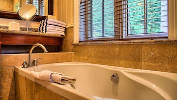 A bathroom with a large bathtub, a mirror, a towel rack, and a window with blinds.
