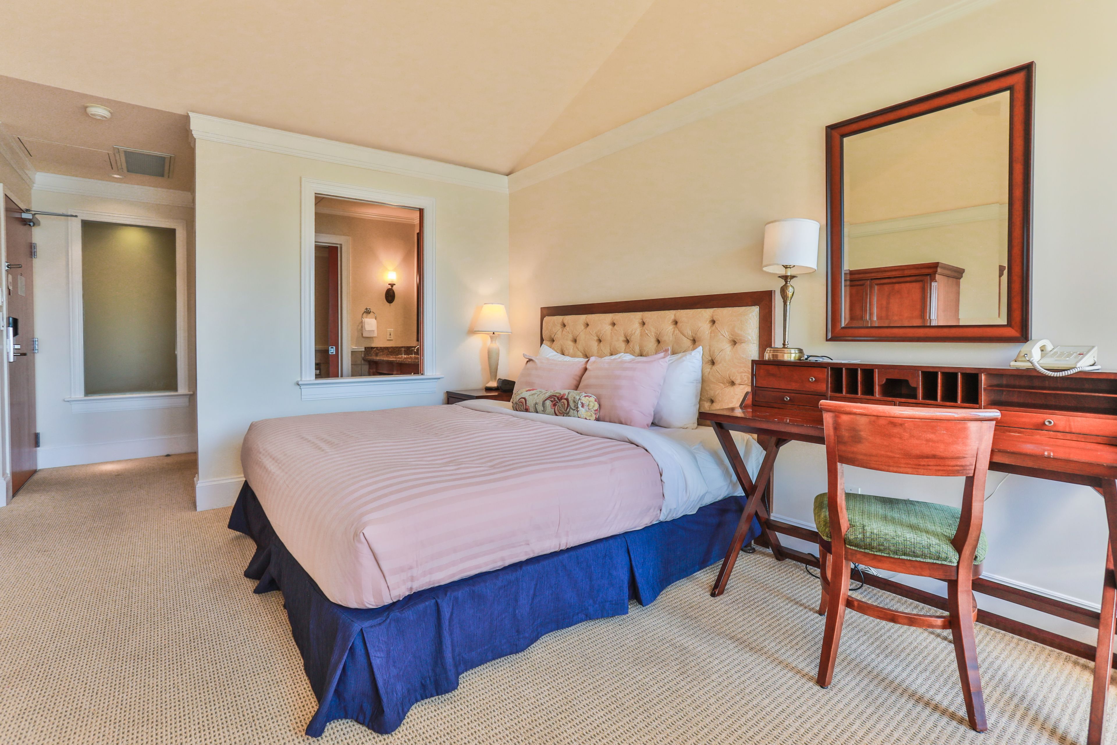 A hotel room with a large bed, a desk with a chair, a mirror, and a lamp.