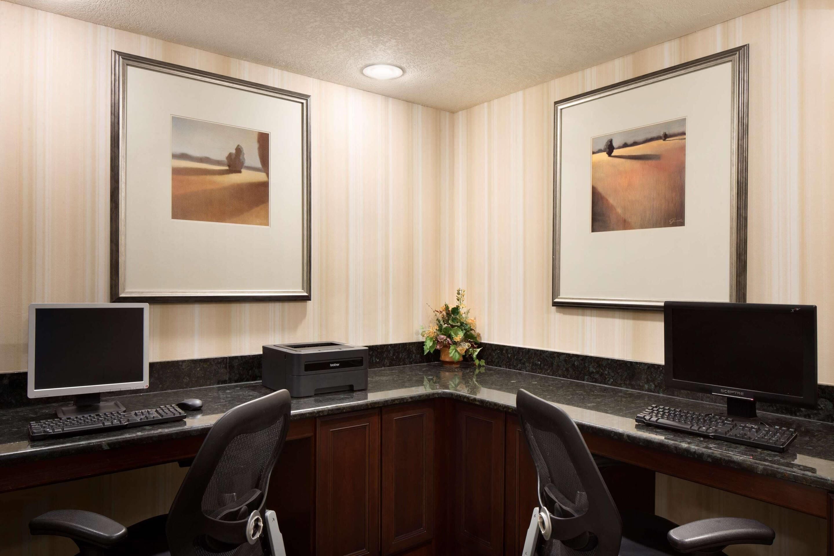 A hotel office with two desks, each with a computer and keyboard, framed artwork on the walls, and a small plant on the counter.