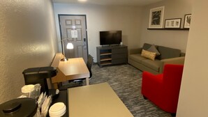 A compact living room with a sofa, a red chair, a TV, and a coffee station. - Country Inn & Suites by Radisson, Champaign North, IL (Champaign)