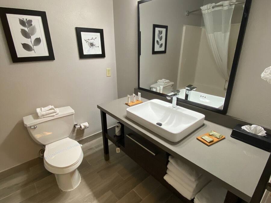 A modern bathroom with a white toilet, a large mirror, and a white sink. There are framed pictures on the wall and a towel rack.