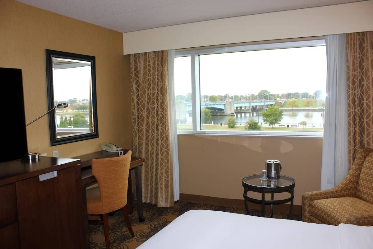 Une chambre d’hôtel équipée d’un lit, d’un bureau, d’une chaise, d’une petite table, d’un téléphone et d’une fenêtre donnant sur un pont et une rivière.