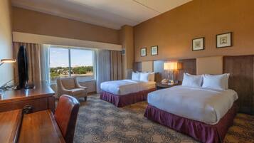 A hotel room with two beds, a desk, a chair, and a large window offering a city view.