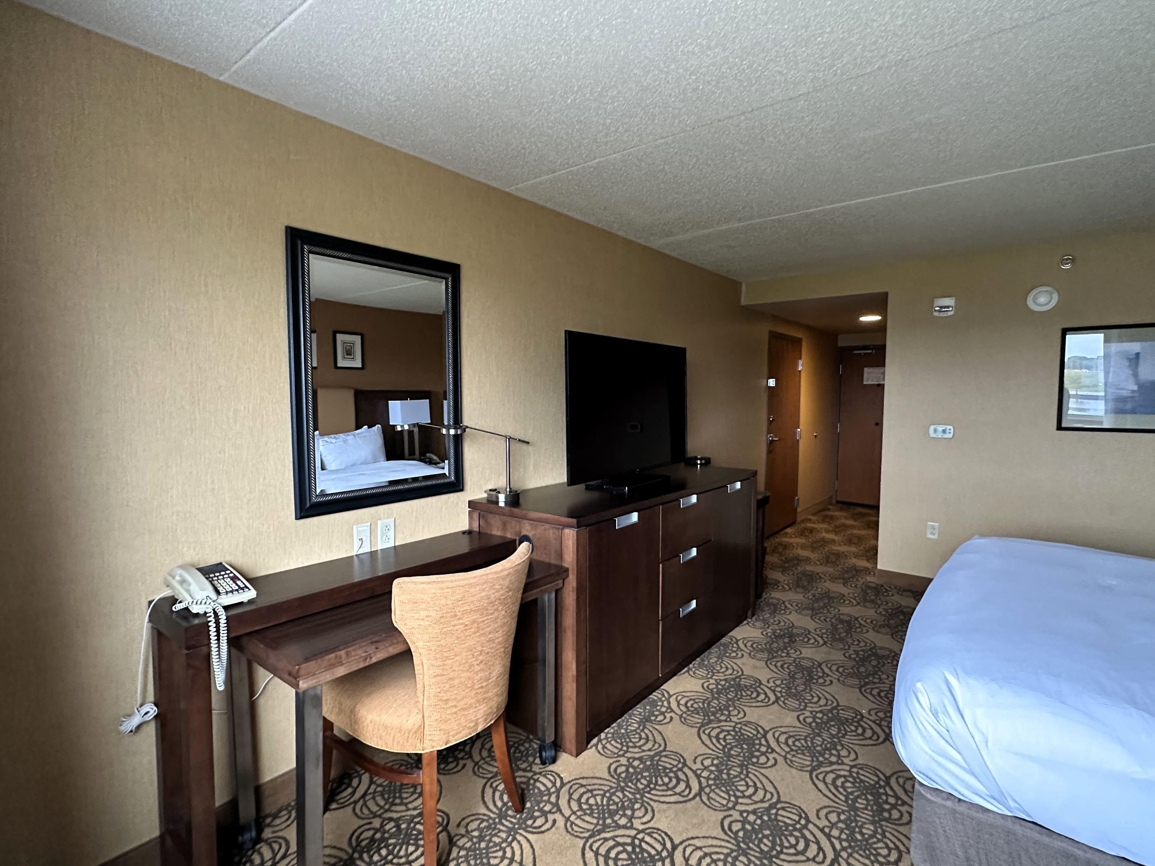 Ein Hotelzimmer mit einem Bett, einem Schreibtisch mit Telefon, einem Sessel, einem Fernseher und einem großen Spiegel.
