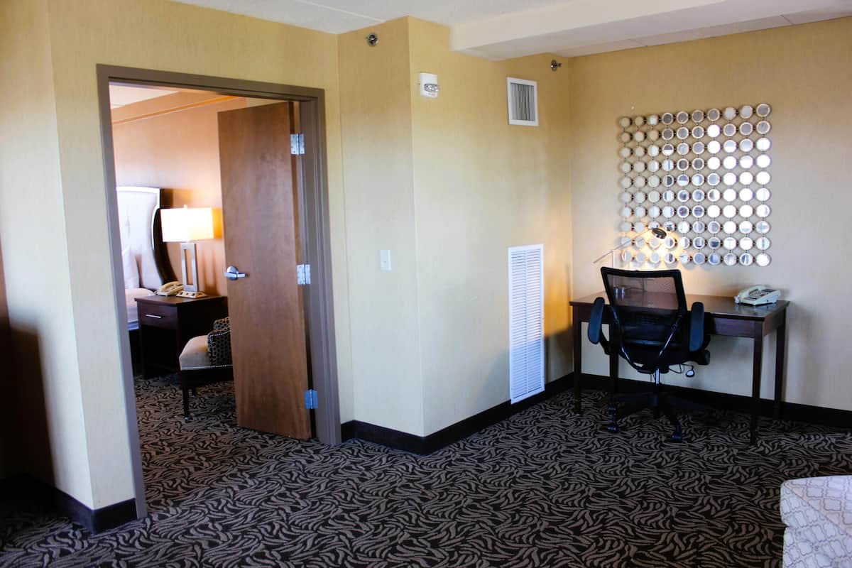 Une chambre d’hôtel avec une moquette à motifs, un bureau avec une chaise, un miroir fixé au mur et une porte donnant sur une autre pièce.
