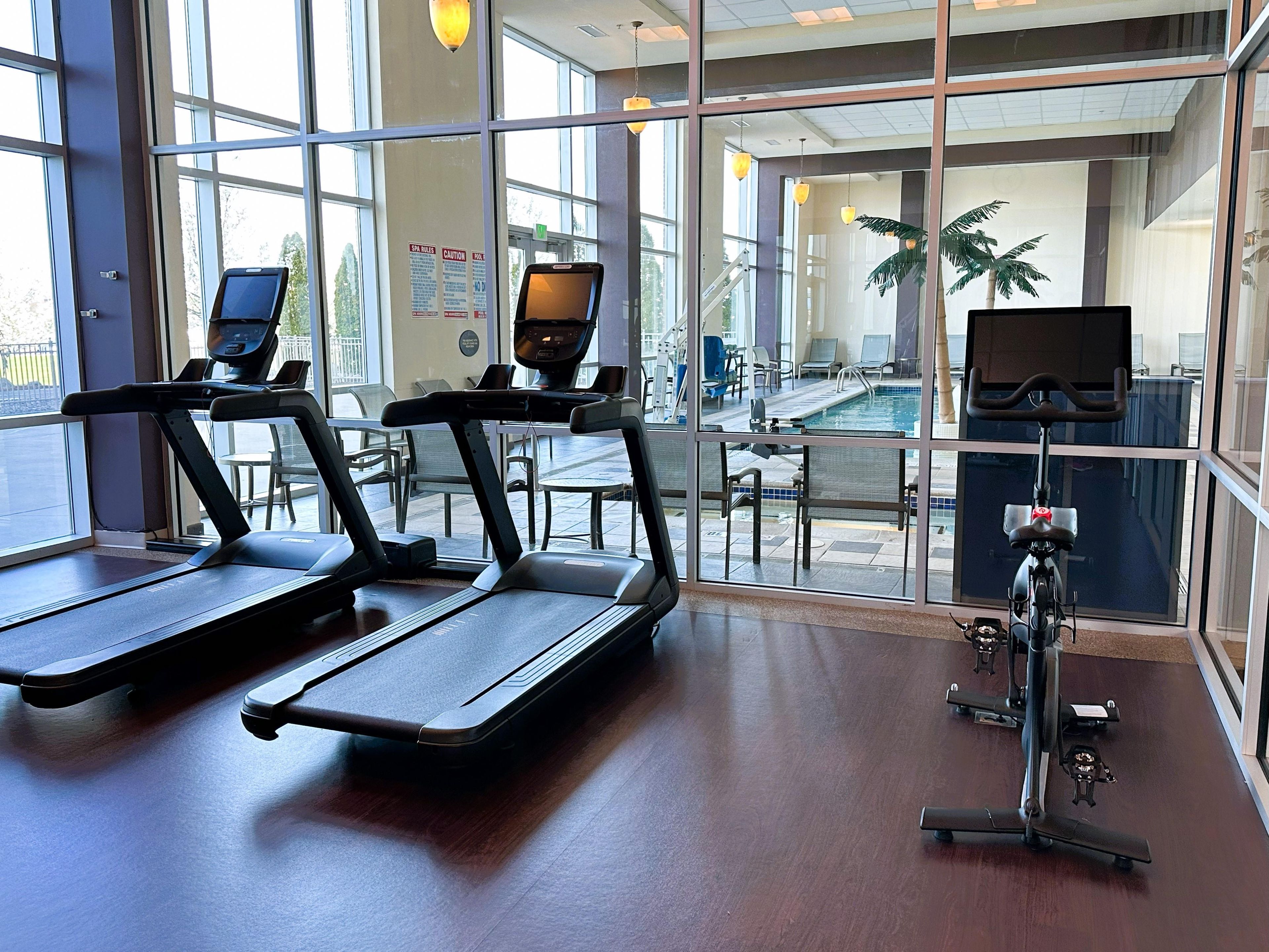 Un gimnasio con cintas de correr, una bicicleta estática y una zona de piscina visibles a través de amplios ventanales.
