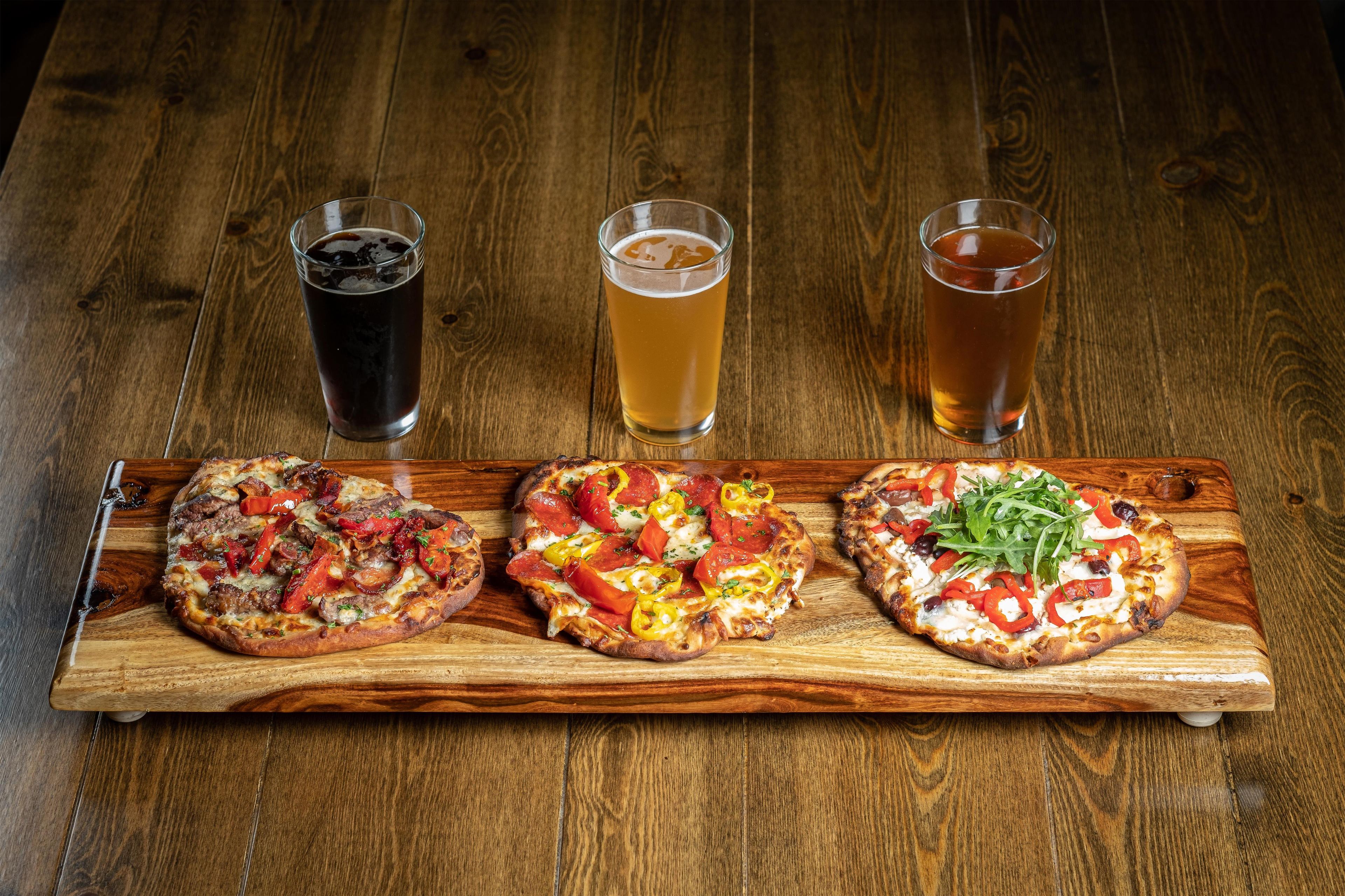 Tres pizzas sobre una tabla de madera con tres vasos de cerveza.