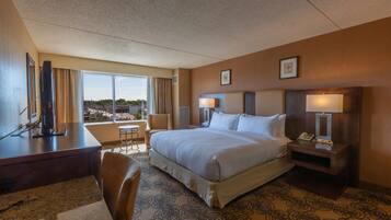 A hotel room with a large bed, a desk, a chair, a small table, and a view of the city.
