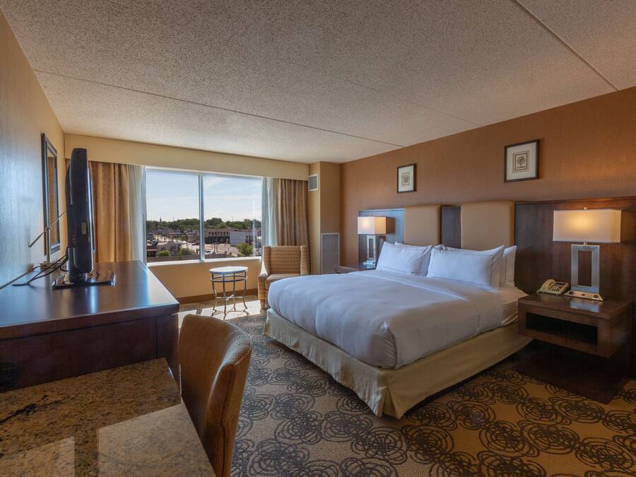 A hotel room with a large bed, a desk, a chair, a small table, and a view of the city.