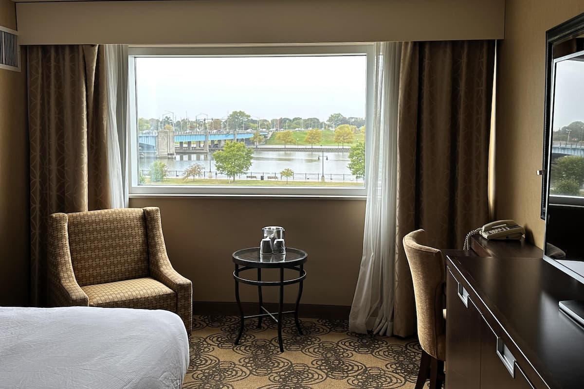 Une chambre d’hôtel avec un lit, une chaise, une petite table, un bureau et une vue sur un pont et une rivière.