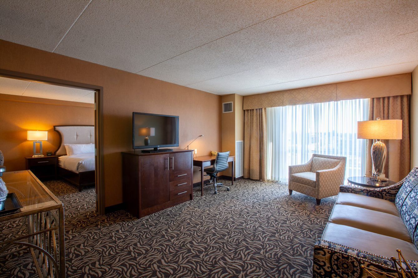Una habitación de hotel con cama, escritorio, televisión, sillón, sofá y ventana con cortinas.