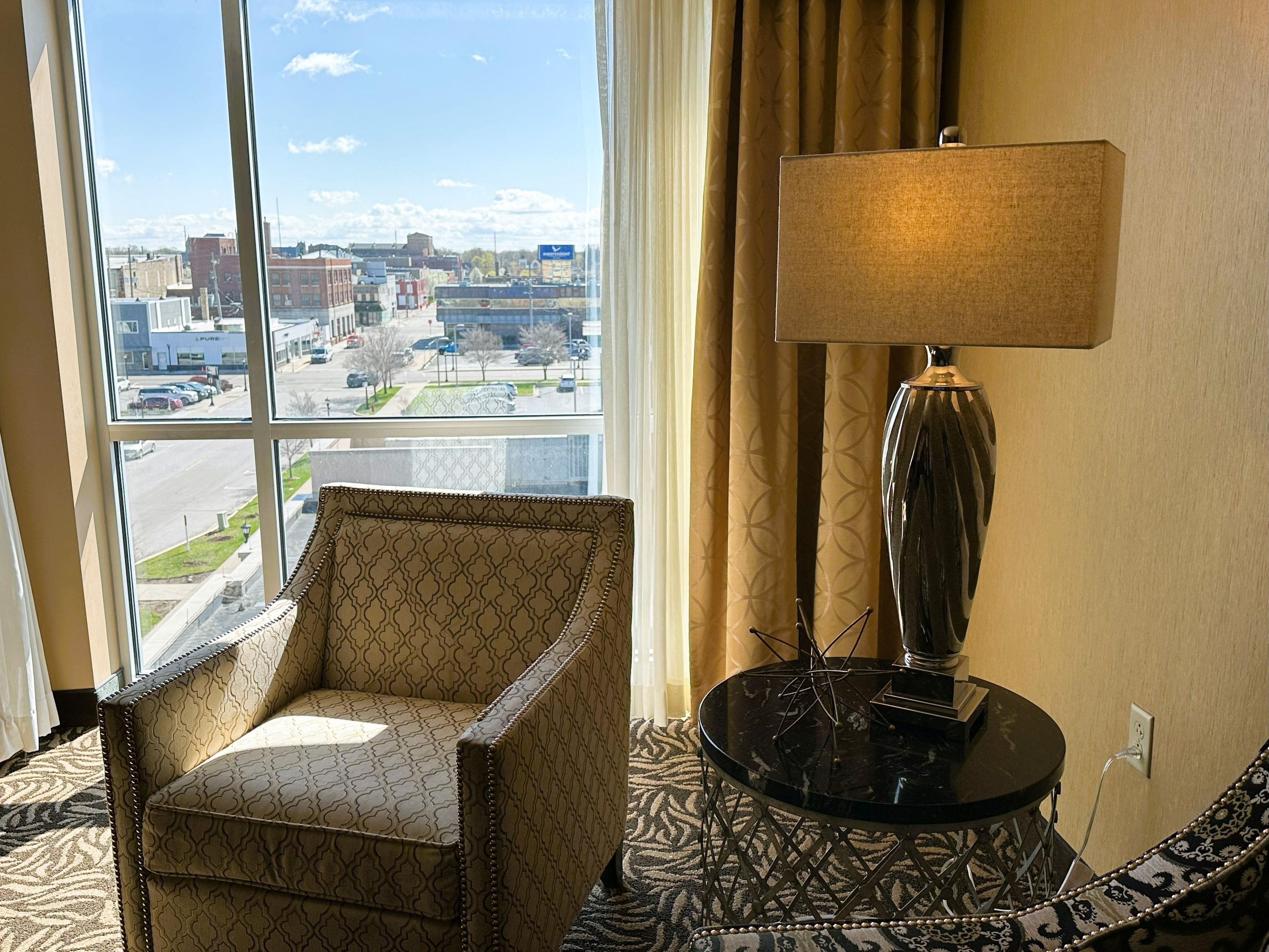 Ein Hotelzimmer mit großem Fenster, einem gemusterten Sessel, einem runden Tisch mit Lampe und einem Teppich mit Muster.