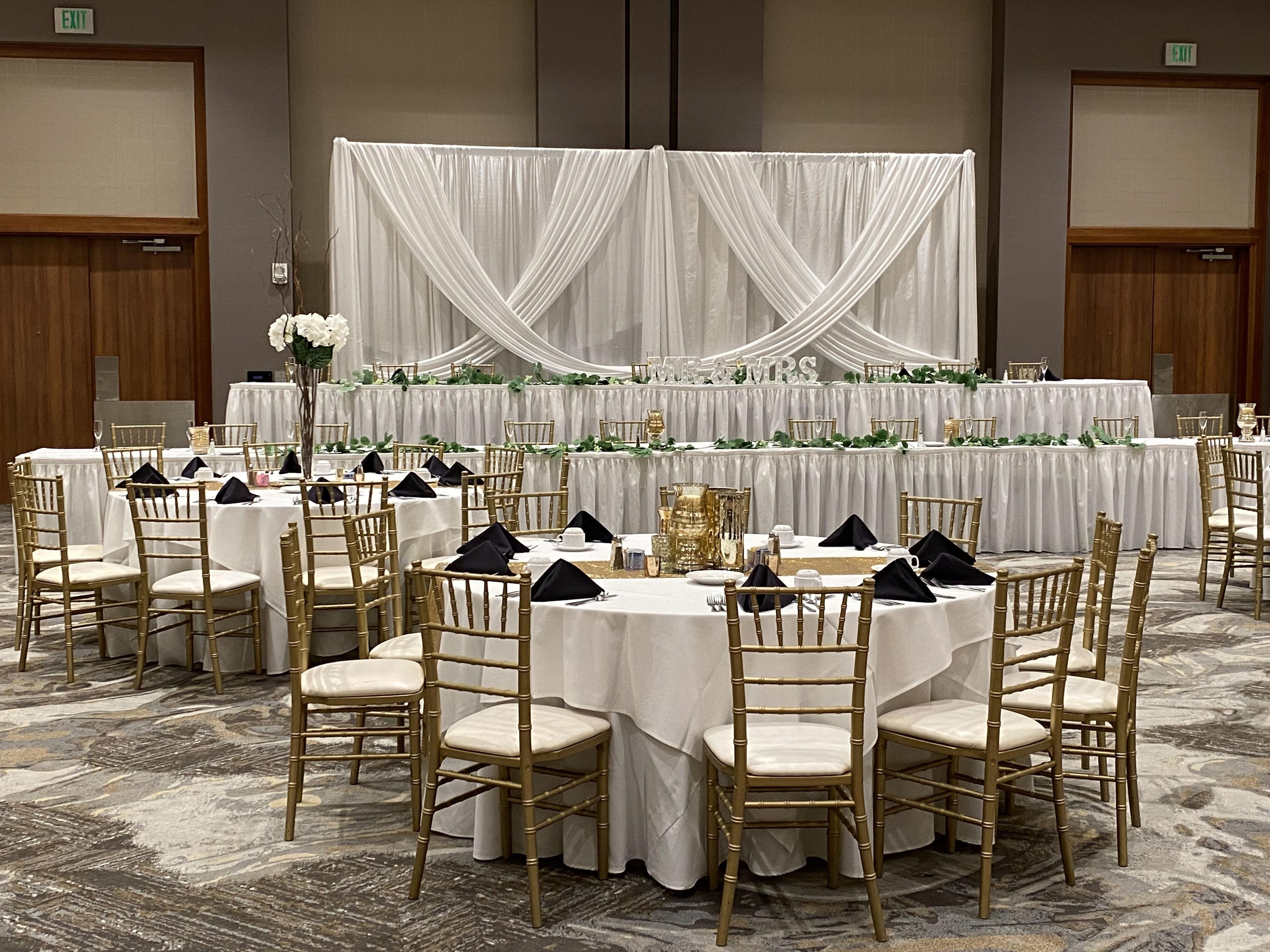 Un salón de banquetes con mesas redondas, sillas doradas, manteles blancos y un telón de fondo con telas drapeadas.