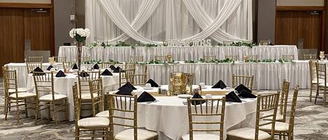 Un salón de banquetes con mesas redondas, sillas doradas, manteles blancos y un telón de fondo con telas drapeadas.