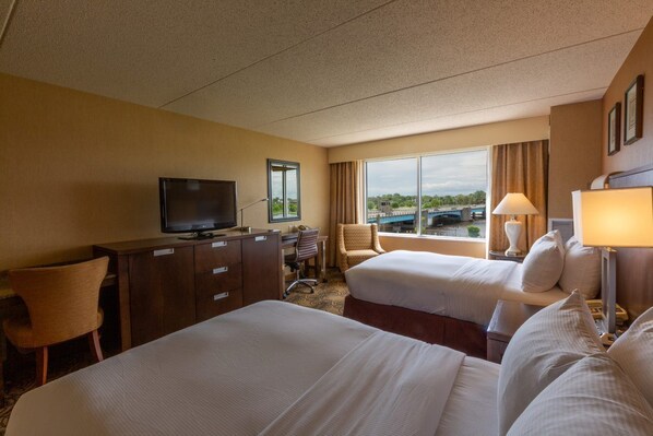 A hotel room with two beds, a desk with a television, a chair, and a window with a view of a bridge. - DoubleTree by Hilton Bay City - Riverfront (Bay City)