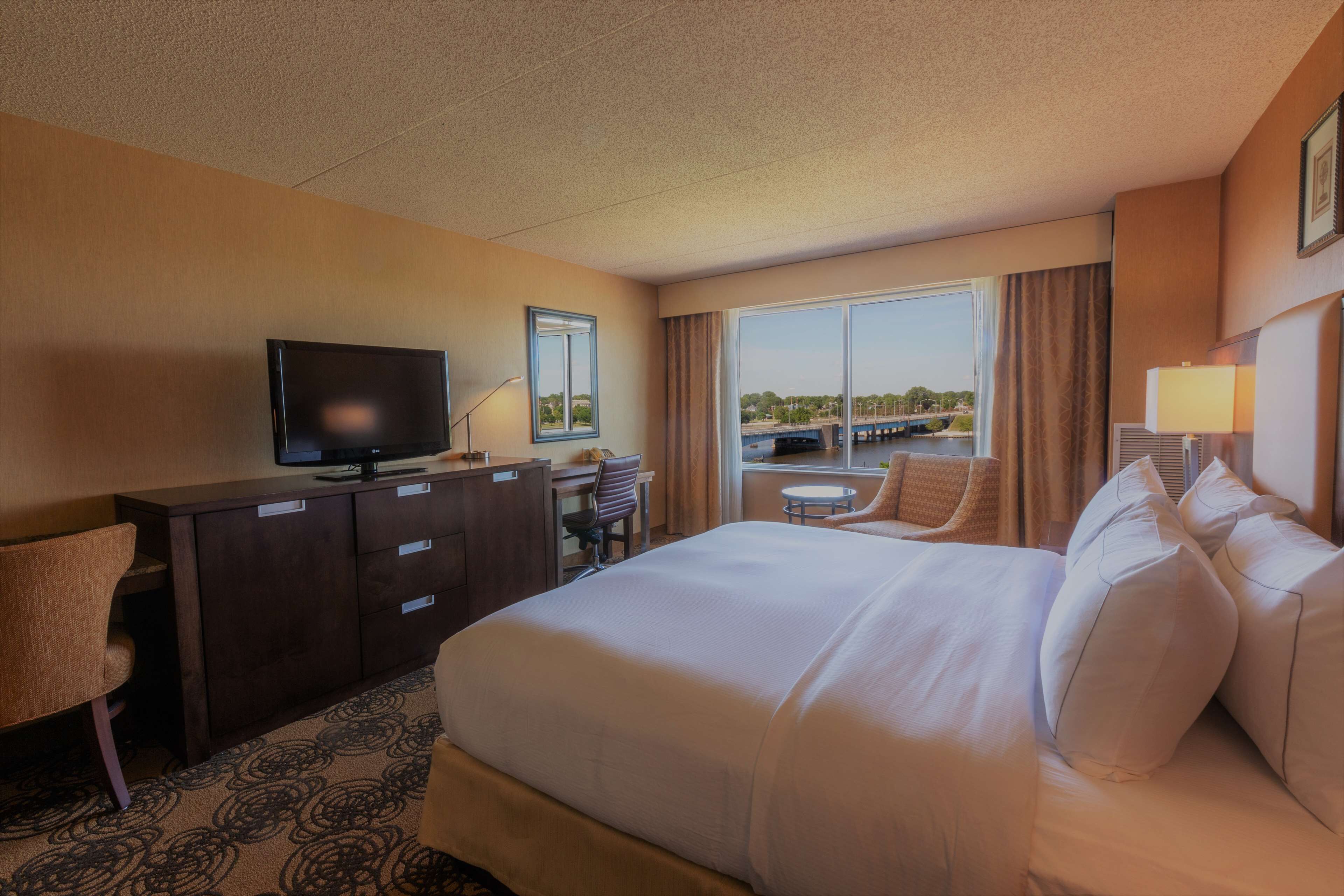 A hotel room with a large bed, a desk with a television, a chair, and a window with a view of a bridge.
