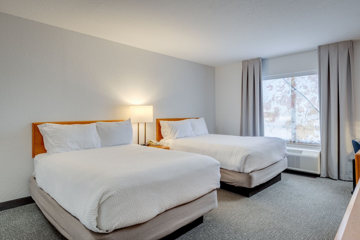 Une chambre d’hôtel avec deux lits, une lampe de chevet, une fenêtre avec des rideaux et une vue sur les arbres à l’extérieur.