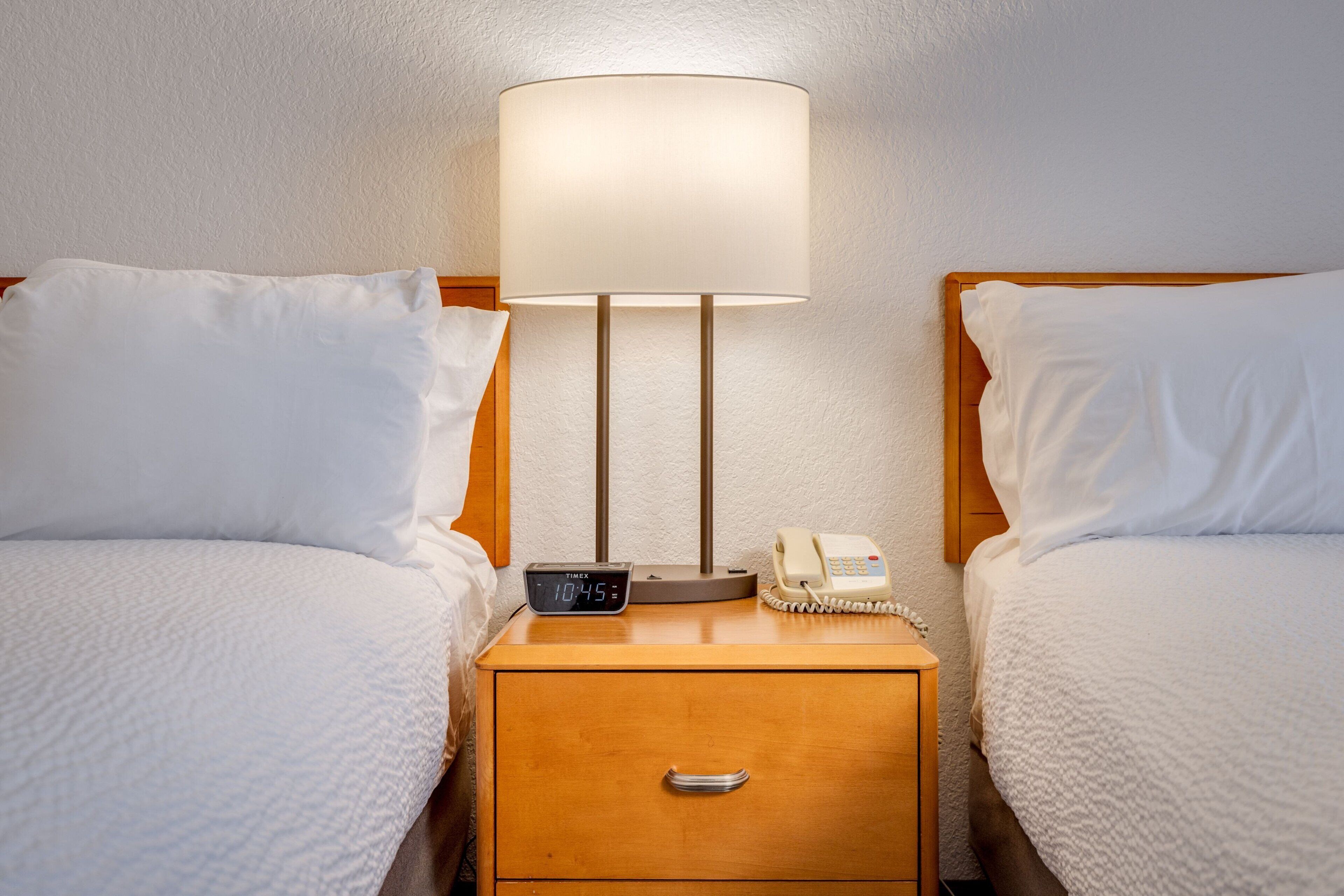 A hotel room with two beds, a bedside table with a lamp, a clock, and a telephone.