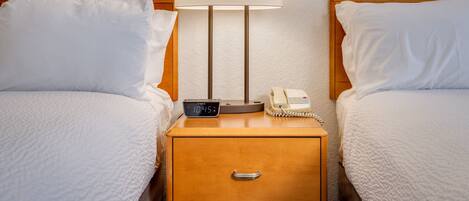 A hotel room with two beds, a bedside table with a lamp, a clock, and a telephone.