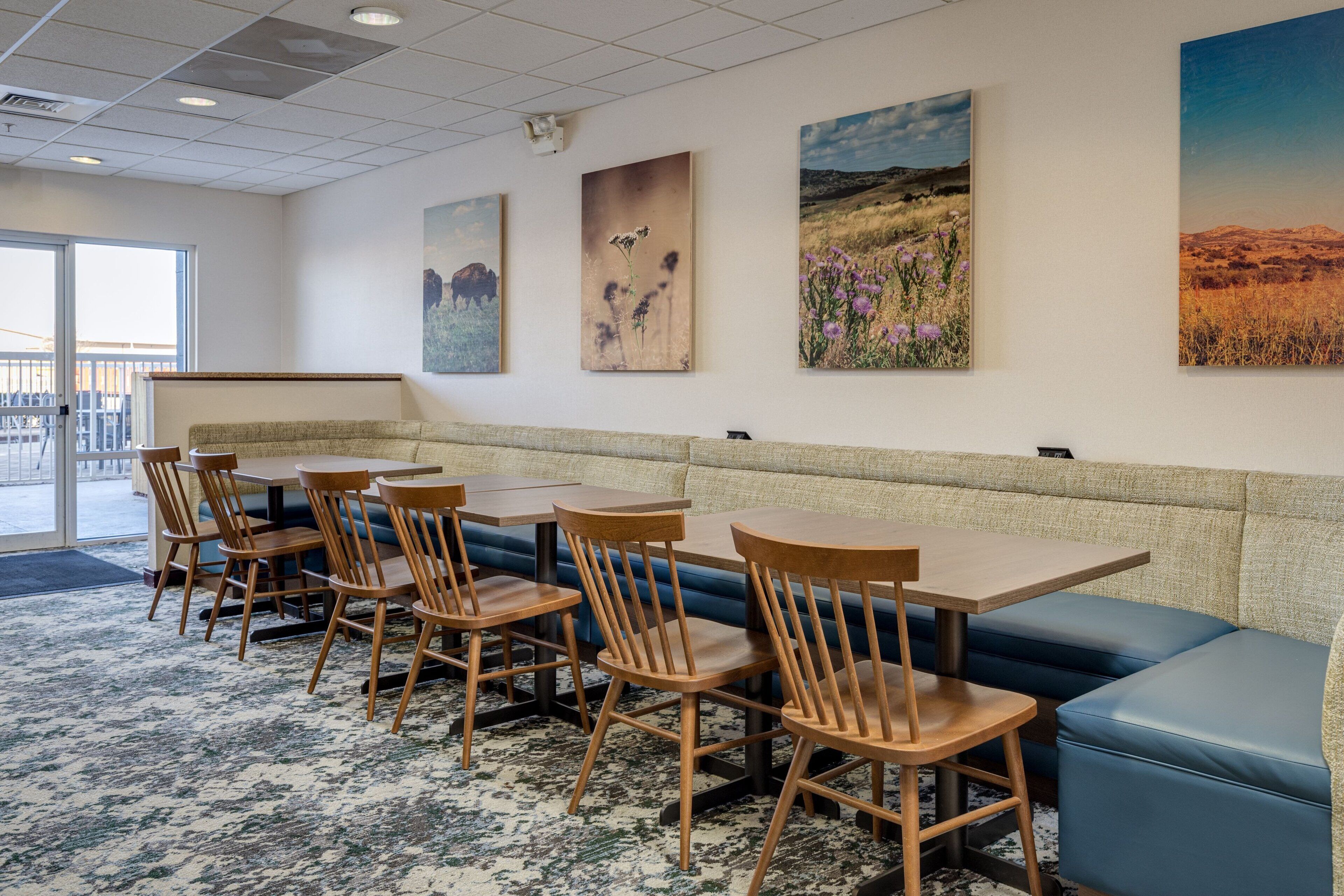 A dining area with wooden tables and chairs, a blue cushioned bench, and four landscape paintings on the wall.