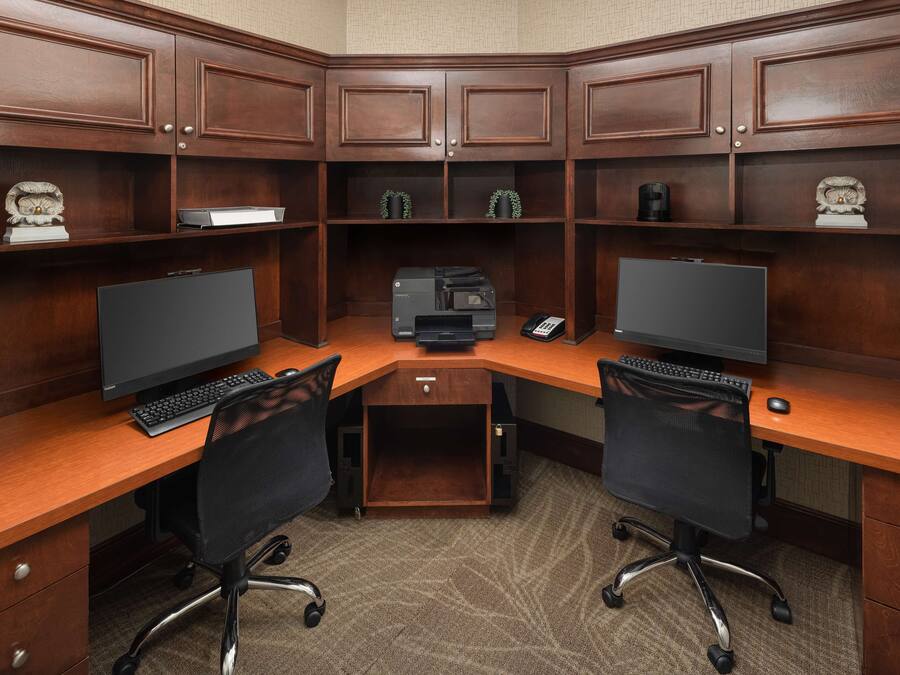 A corner office with two desks, each with a computer, keyboard, and chair. There are shelves above the desks with decorative items and a printer in the center.