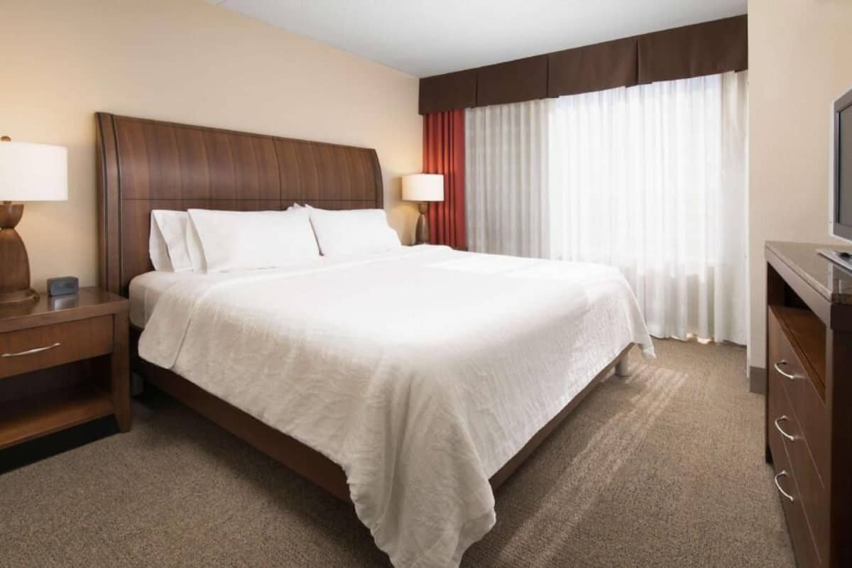 A hotel room with a large bed, two bedside tables with lamps, a TV, and a window with curtains.