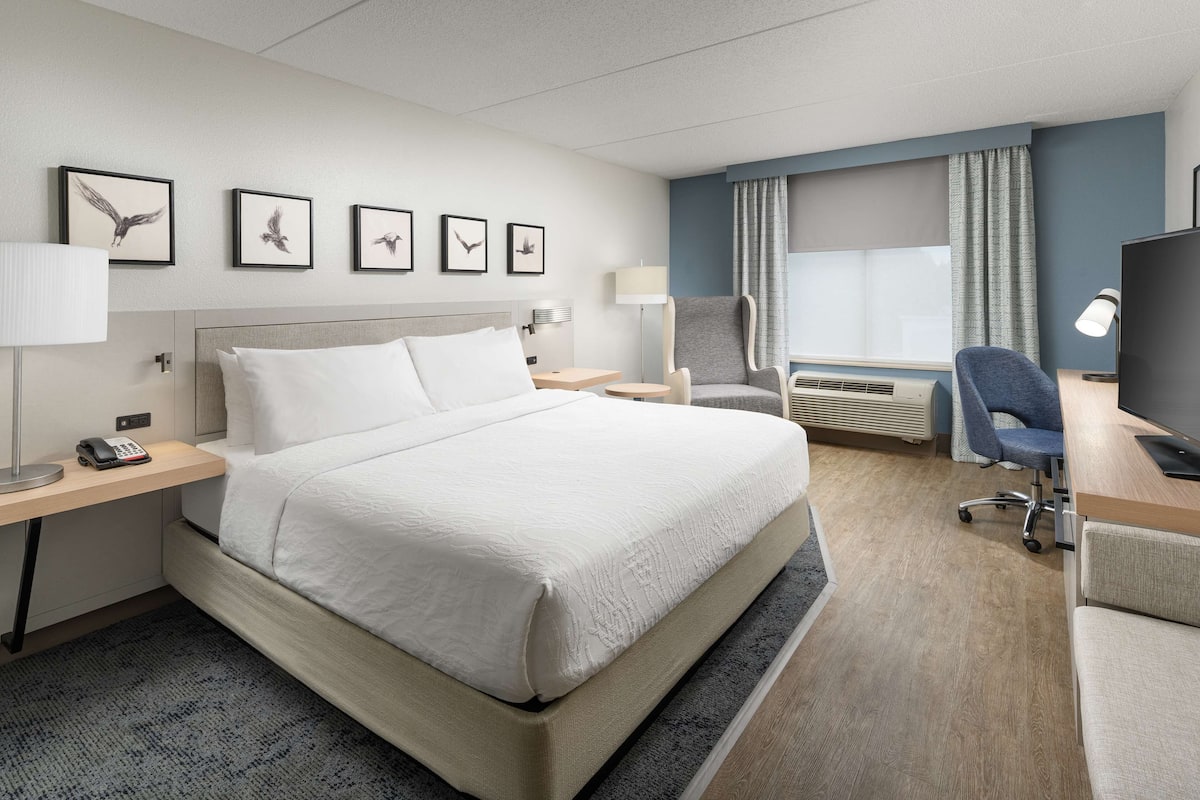 A hotel room with a large bed, a desk with a computer, a chair, and a TV. There are framed pictures on the wall and a window with curtains.