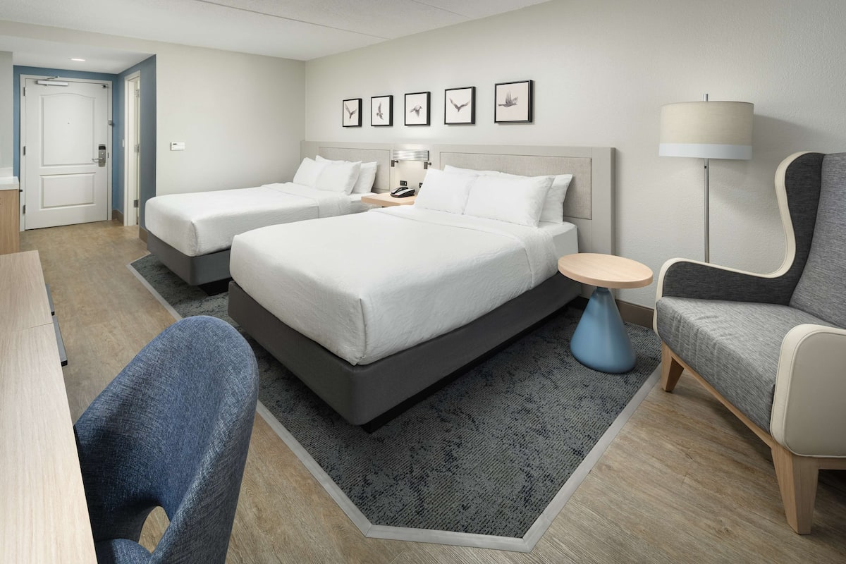 A hotel room with two beds, a desk, a chair, a lamp, and a small table.