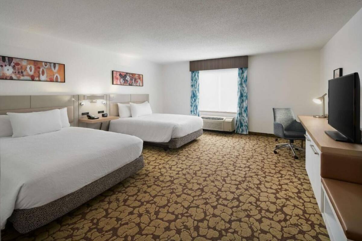 A hotel room with two beds, a desk with a TV, a chair, and a window with curtains.