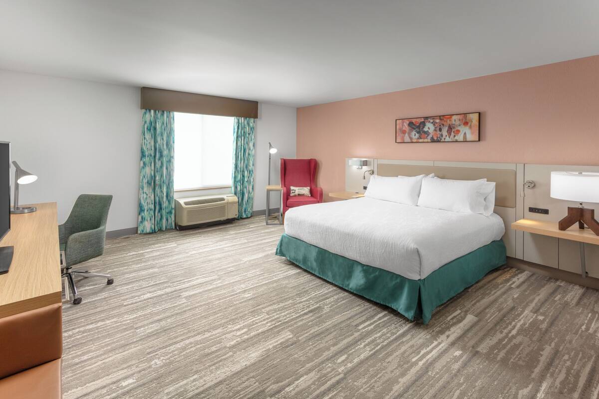 A hotel room with a bed, a desk, a chair, a desk lamp, a window with curtains, an air conditioning unit, and a painting on the wall.