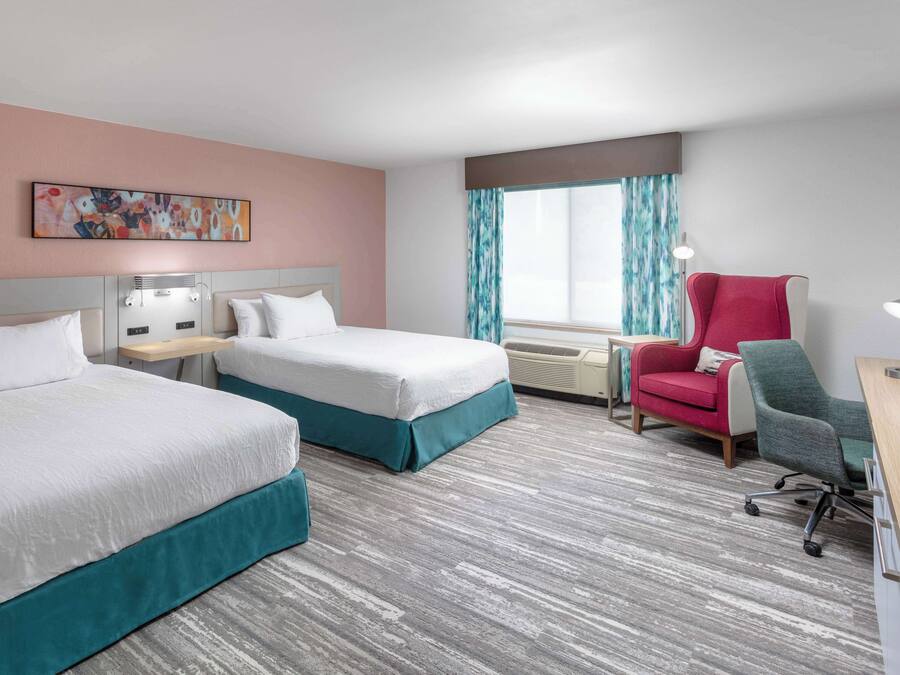 Habitación de hotel con dos camas, un escritorio, una silla y un ventanal con cortinas.