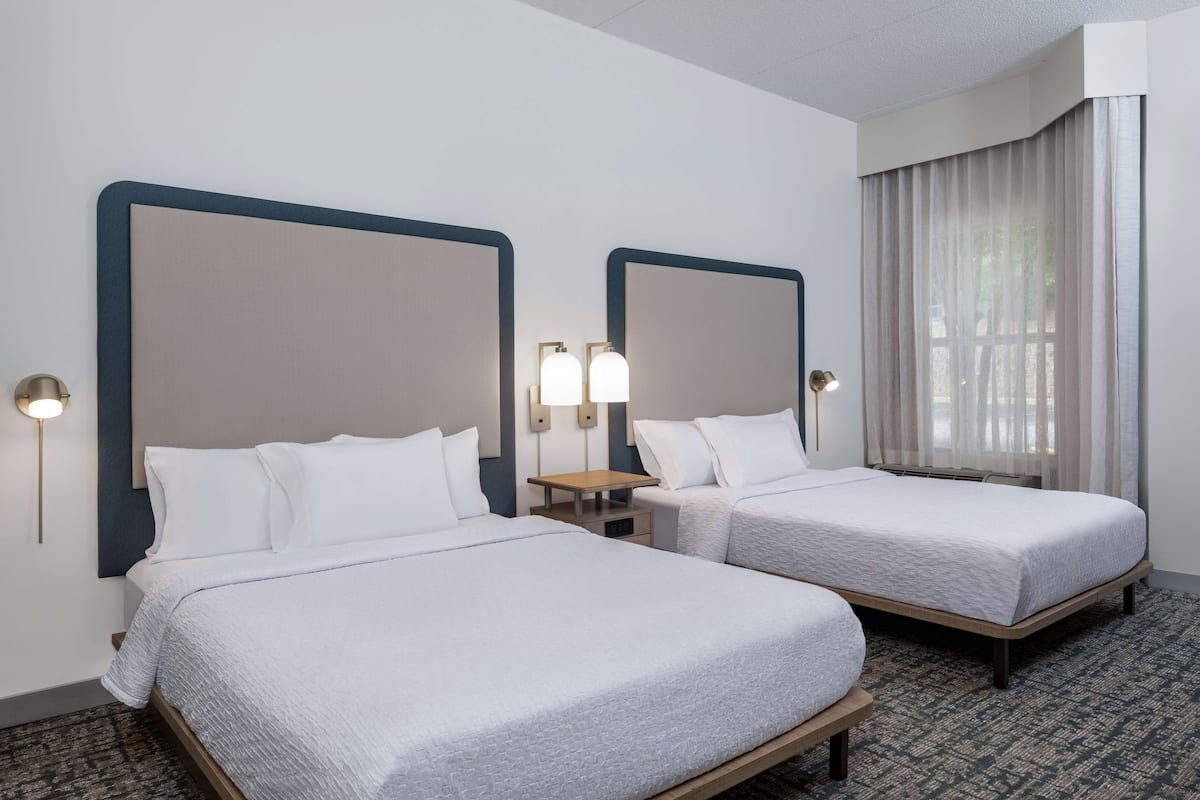 A hotel room with two single beds, a small table, two wall-mounted lamps, and a window with curtains.