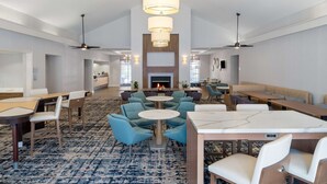A spacious lobby with a central fireplace, multiple seating areas including round tables and chairs, and ceiling fans. - Homewood Suites by Hilton Montgomery (Montgomery)