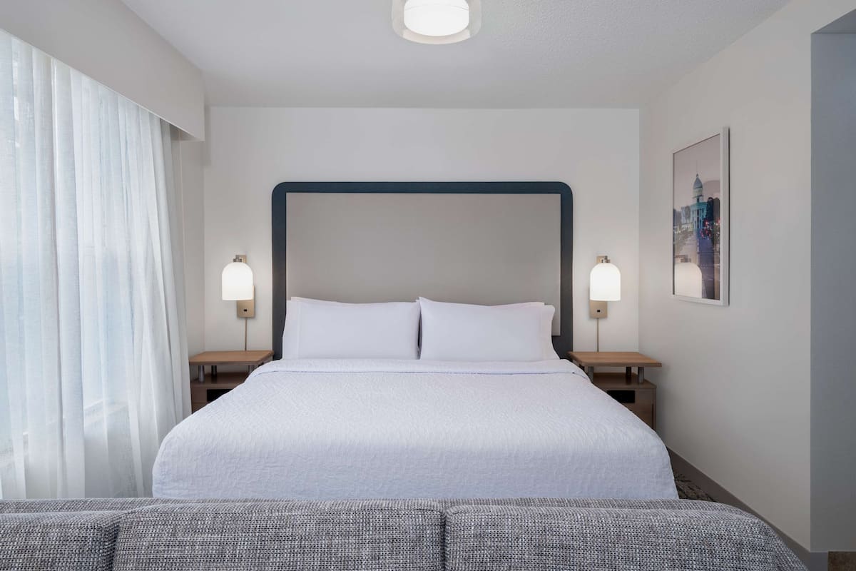 A hotel room with a large bed, two bedside tables with lamps, a window with sheer curtains, and a framed picture on the wall.