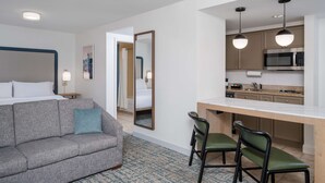 A hotel room with a grey sofa, a small dining table with two chairs, a kitchen area with a microwave, and a large mirror. - Homewood Suites by Hilton Montgomery (Montgomery)