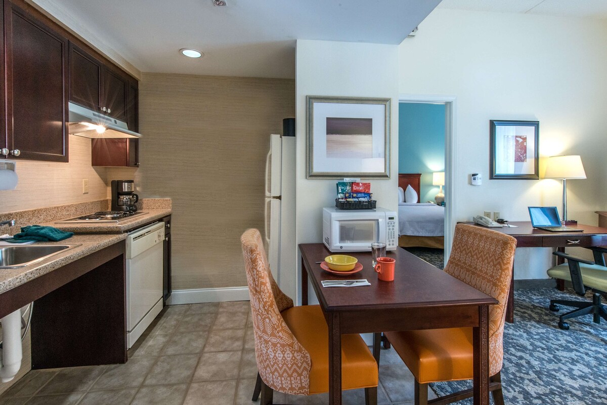 Quarto de hotel compacto com cozinha americana, área de refeições com duas cadeiras, uma cama, escrivaninha com um laptop e uma mesinha com uma tigela e caneca.