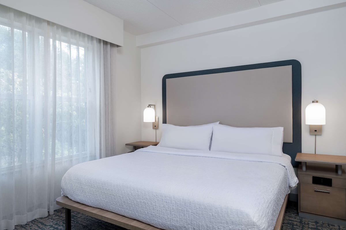 A hotel room with a large bed, two bedside tables, a nightstand, a window with sheer curtains, and wall-mounted lamps.