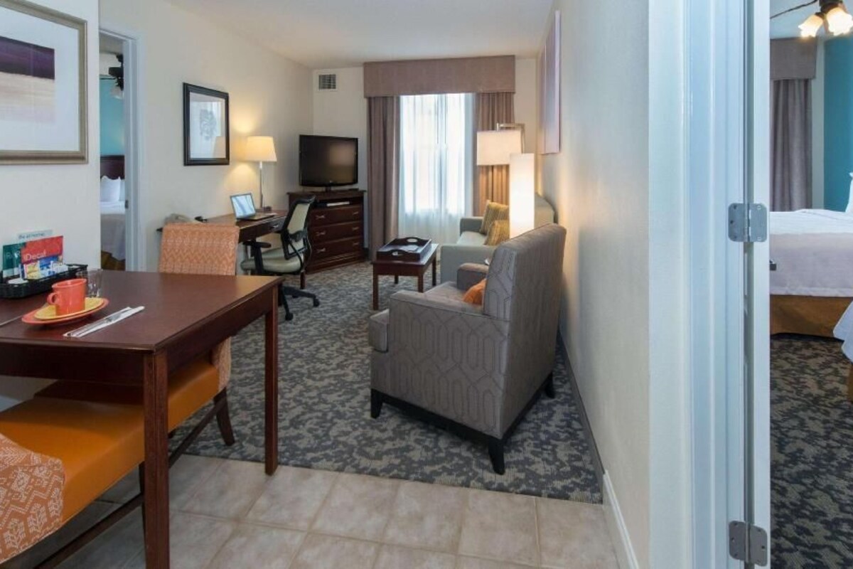 Quarto de hotel com cama, escrivaninha com cadeira, sofá, televisão e abajur.