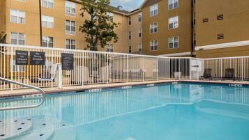 A swimming pool with a white fence, signs indicating no diving and lifeguard duty, and a building with multiple windows and balconies in the background.