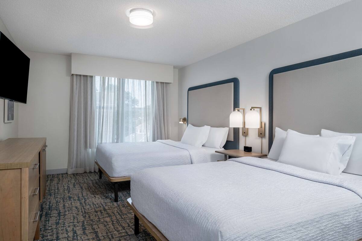 A hotel room with two beds, a desk, a TV, and a window with curtains.