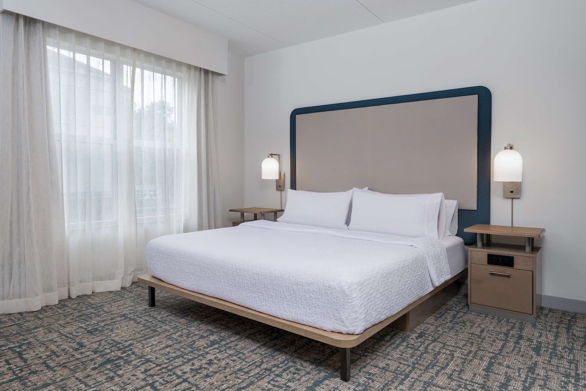 A hotel room with a large bed, two bedside tables, a nightstand, and a window with sheer curtains.