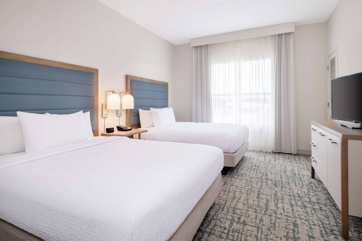 A hotel room with two beds, a desk with a TV, a lamp, and a window with curtains.